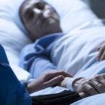 Person lying in hospital bed with oxygen tubes in nose and another person resting their hand on their arm